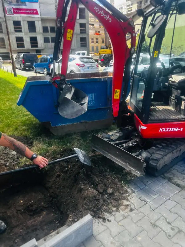 Minikoparka nabierająca ziemię łyżką i ładująca ją do niebieskiego kontenera na urobek.